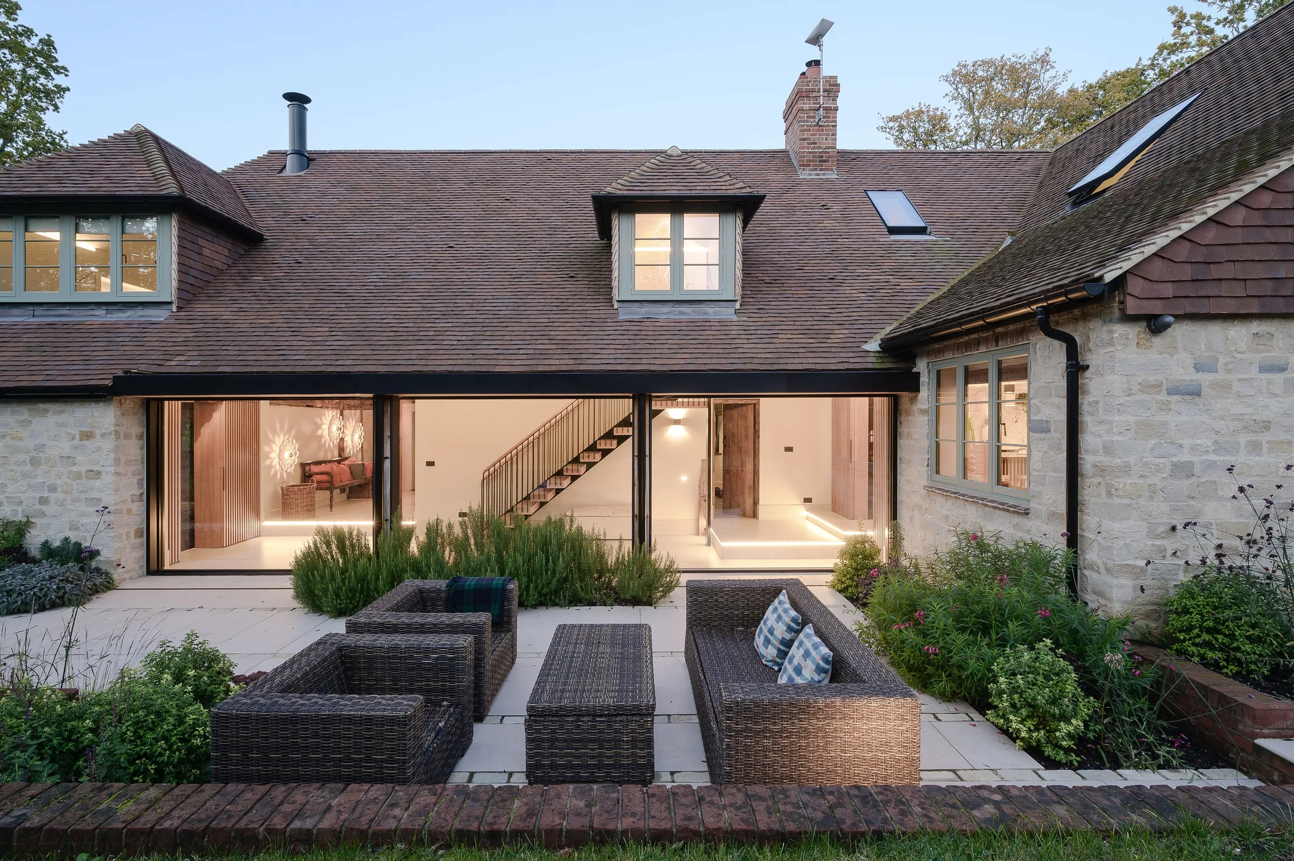 Tiled roof and stone elevation at dusk, sliding doors open above wicker terrace seating, country house, Lodsworth