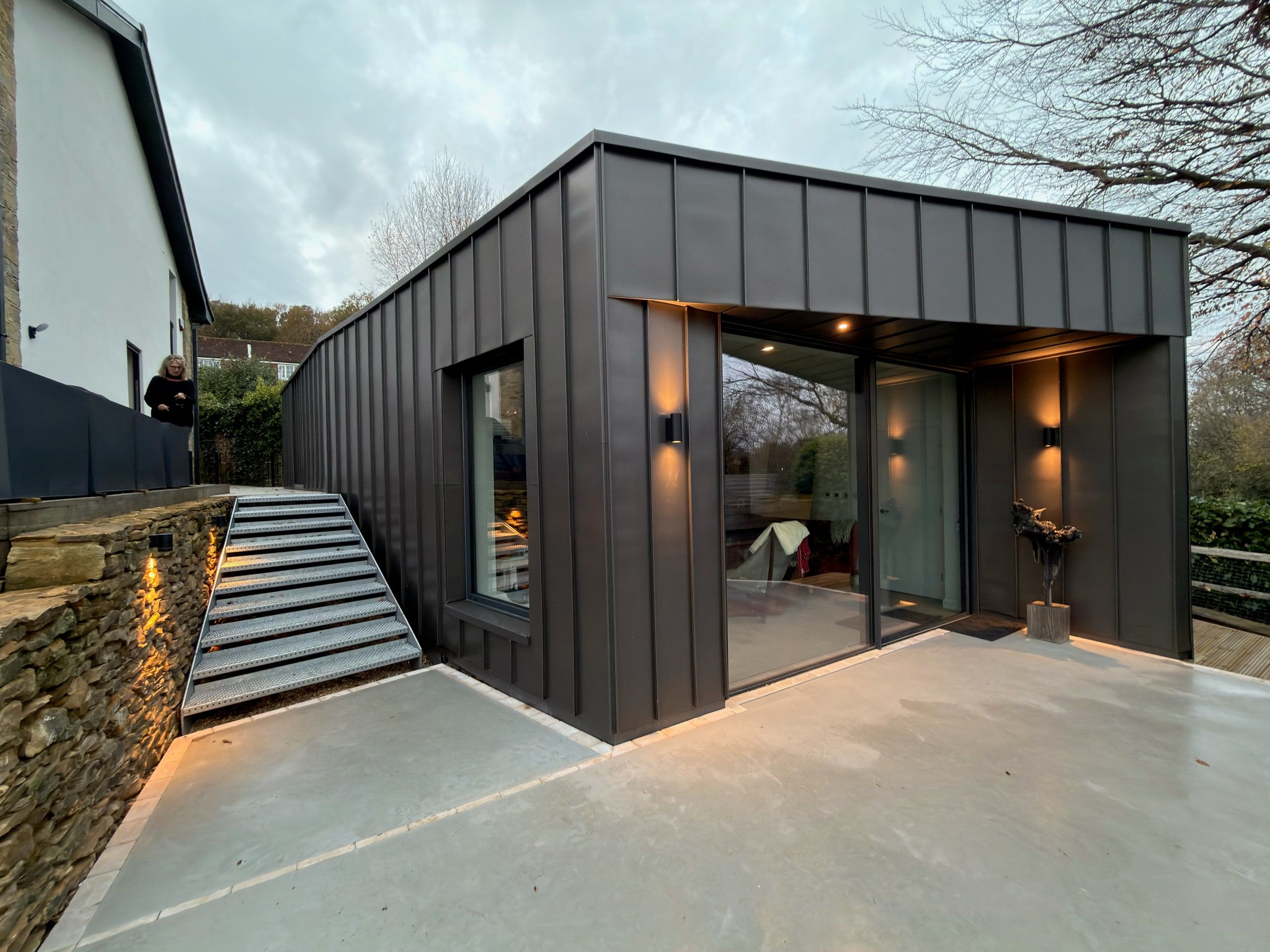 Zinc-clad bay extension lit at dusk above the stone terrace steps, 1960s house, Lodsworth.