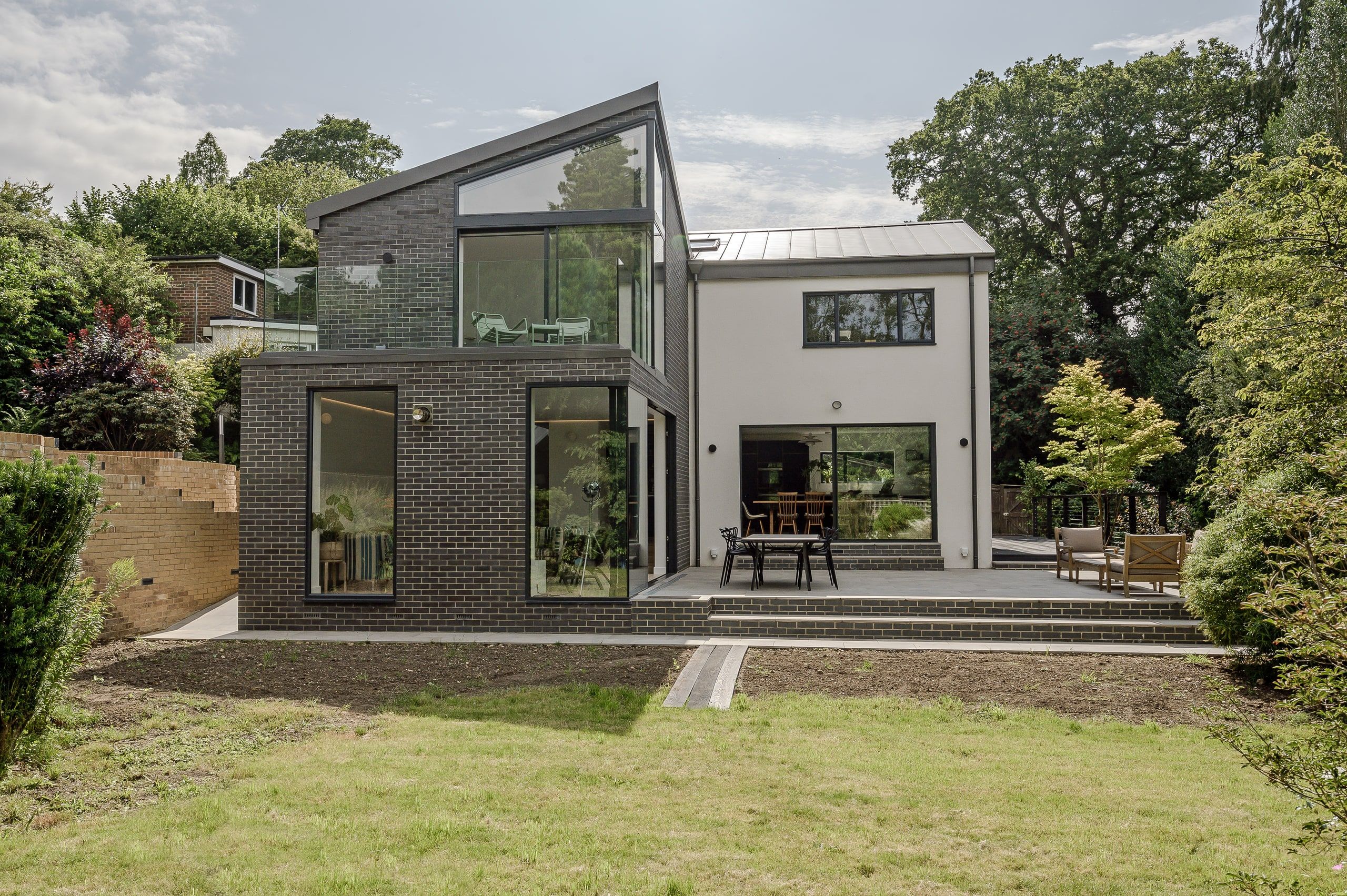 Dark brick extension with a glass balcony beside the rendered house, seen from the garden, 1960s house, Haslemere