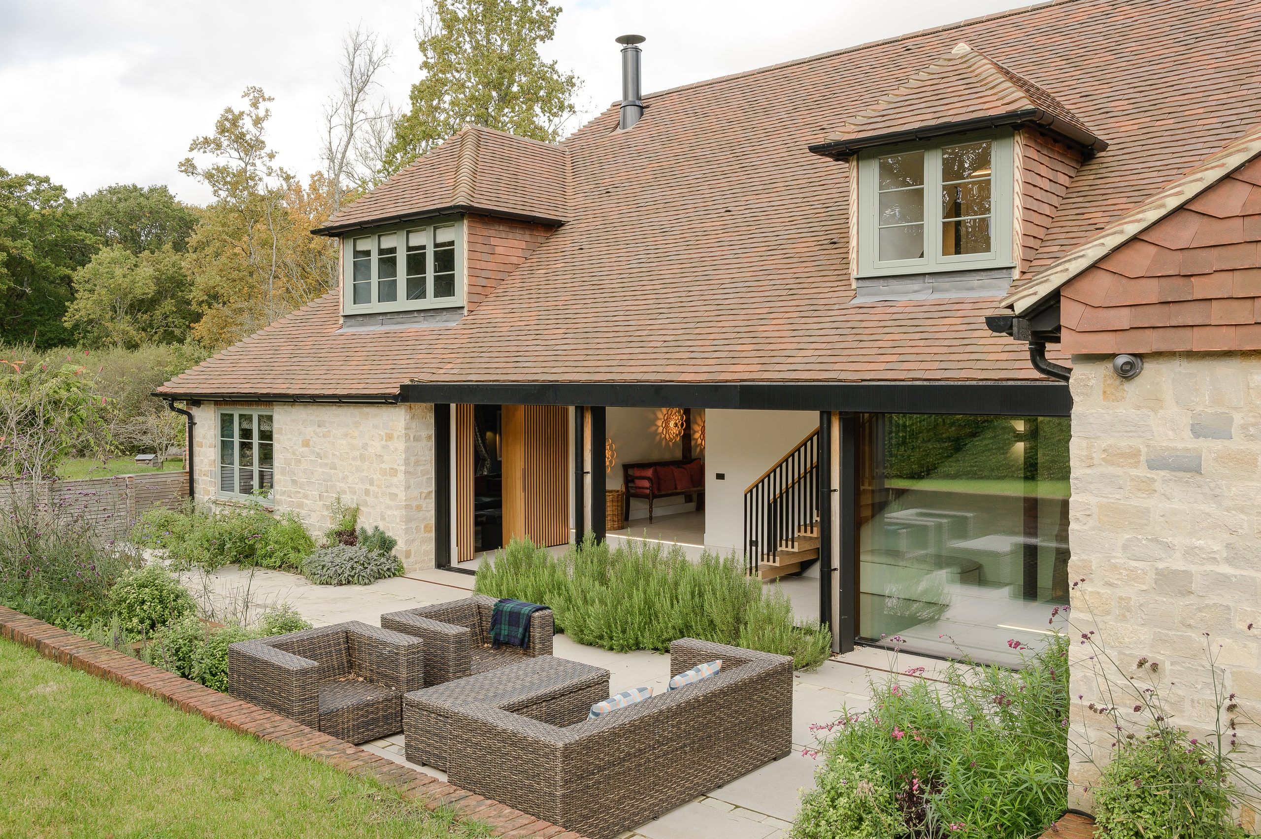 Stone elevation with green-framed dormers, sliding doors open above rattan seating, country house, Lodsworth