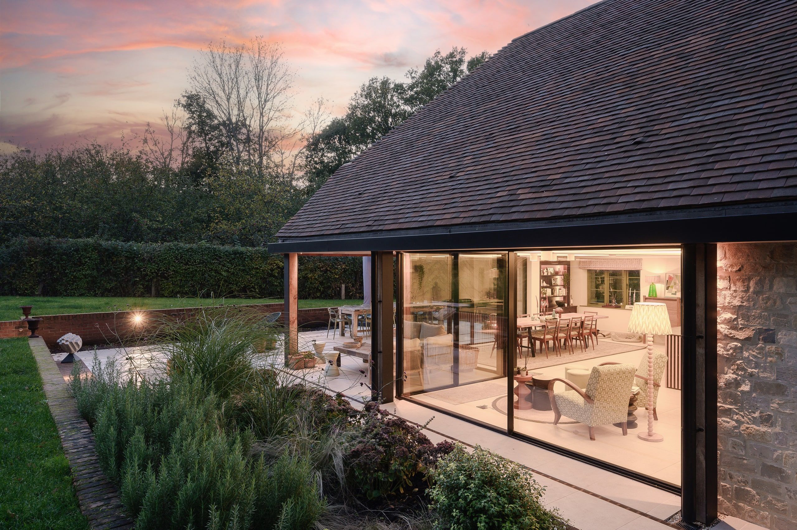 Sunset view along the glazed dining room, lamplight inside and herb planting outside, country house, Lodsworth